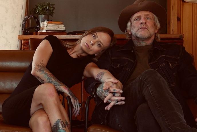 Tattooed artist duo sits close on couch in warm wood room, posing for a picture.