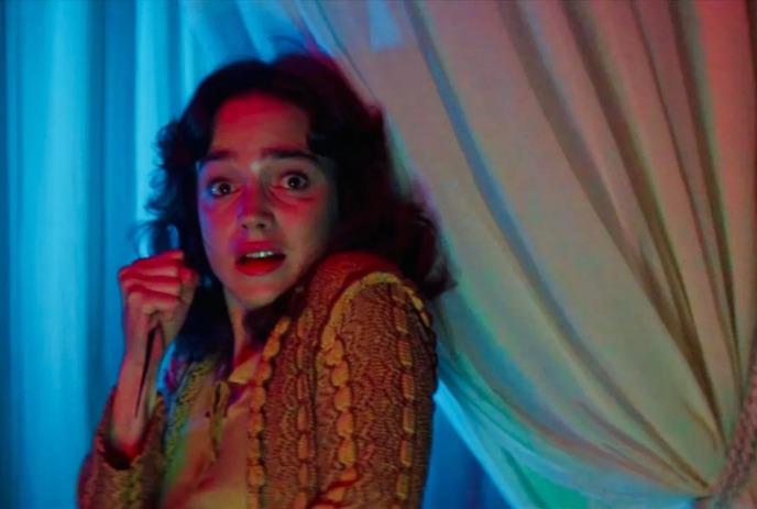 Person in yellow-brown garment stands before glowing curtains, clenched hand raised in fear, lit by red and blue tones.