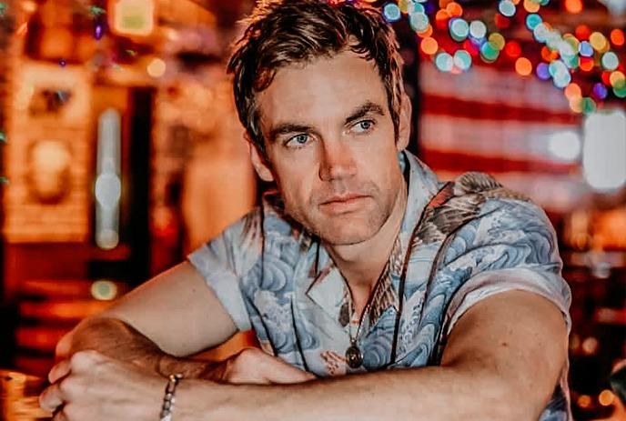 Tyler Hilton seated indoors with colorful lights in the background.