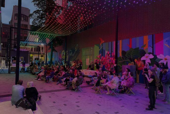 People gather under colorful lights and a mural, enjoying an outdoor urban event with lively ambiance.