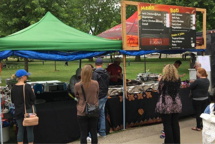 A food stall with a green canopy offers meals and roti at an outdoor event, as attendees wait in a queue.