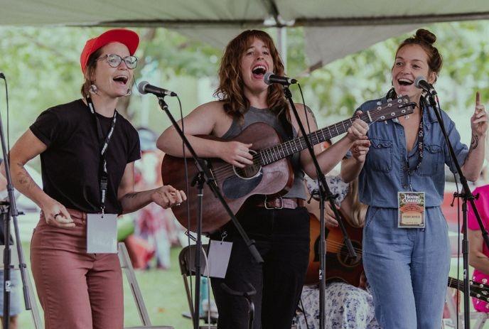 Three performers on stage: one playing guitar, two singing into microphones, under a tent and green background.