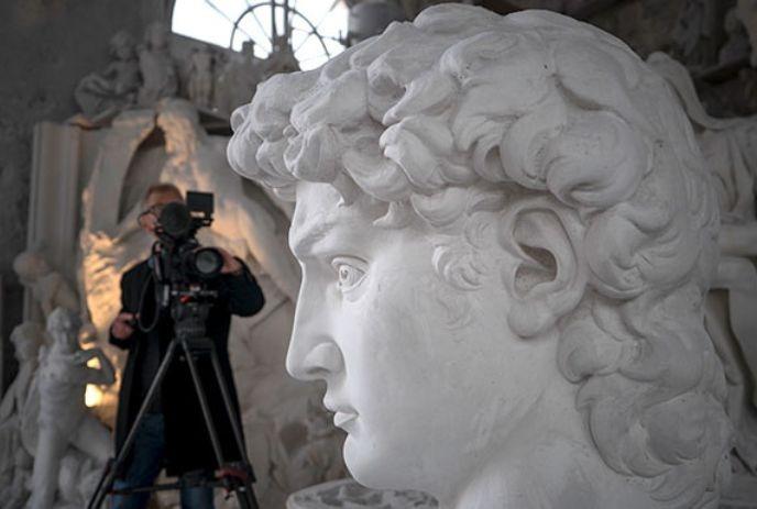 An artist films a large, intricate head sculpture in a studio with other sculptures and natural light.