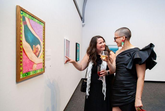 Two womans in black party dresses checking an art piece while holding a glass of wine.