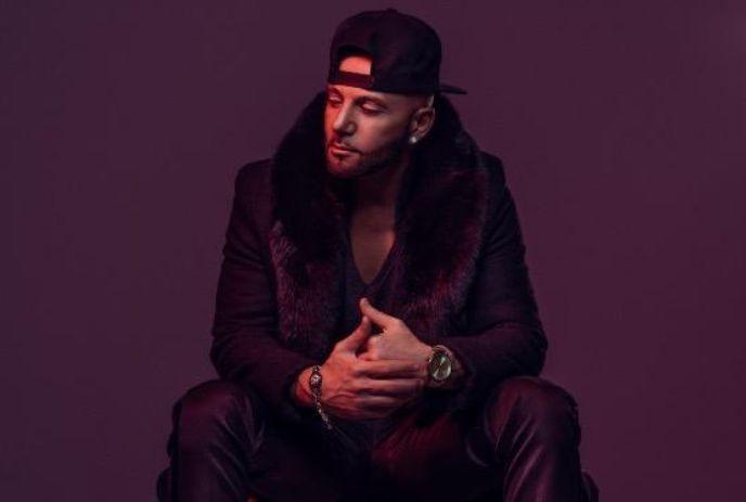 Karl Wolf sitting under a dark background with black outfit and a hat posing for a picture in a dim atmosphere.