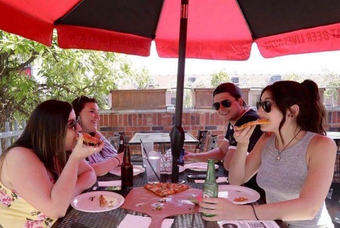 A group of four people enjoying pizza and drinks under a red umbrella at an outdoor table.