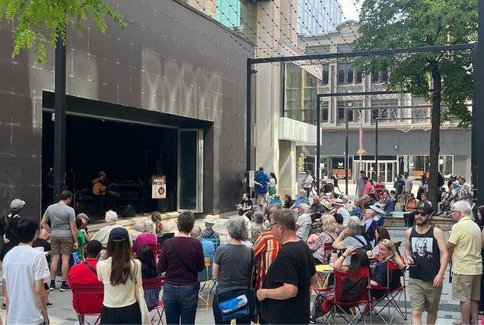 Outdoor concert in city; crowd watches musicians perform on stage, seated and standing attendees enjoying live music.