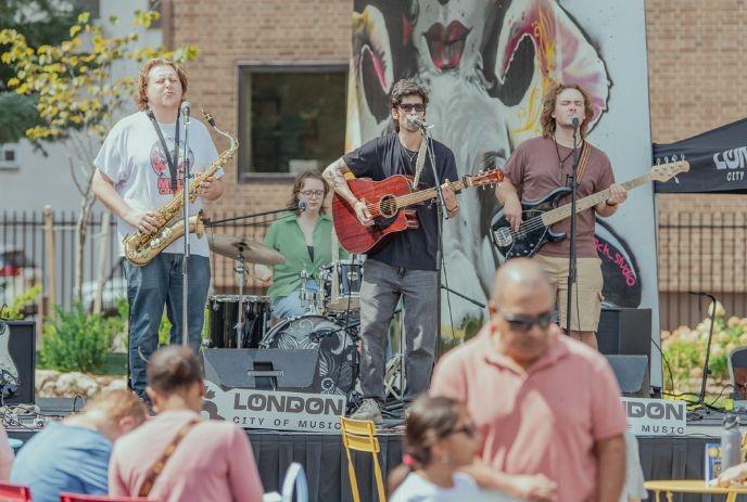 A band performs on an outdoor stage. The backdrop features a mural with 'London City of Music' on the stage.
