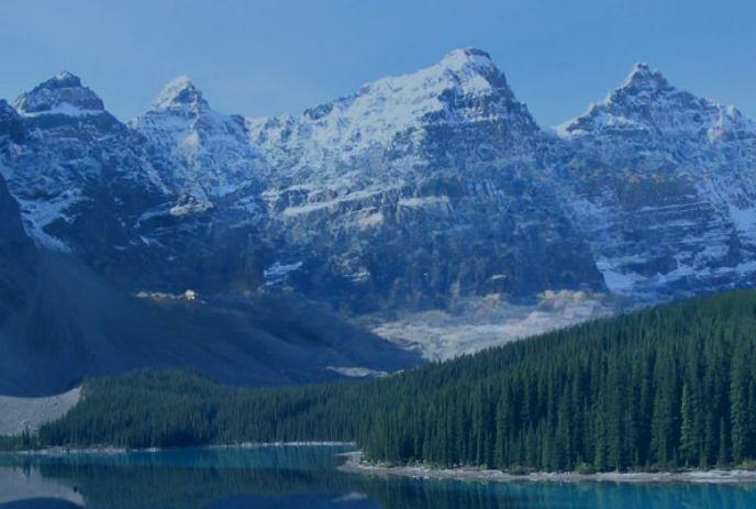 Snowy peaks, rugged mountains, lush forest, and a reflective lake under a bright blue sky