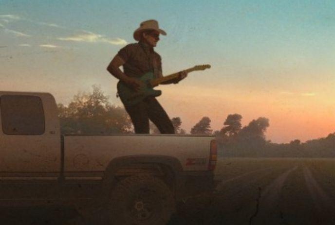 Brad Paisley wearing a cowboy hat stands in the back of a pickup truck, playing an electric guitar at sunset.
