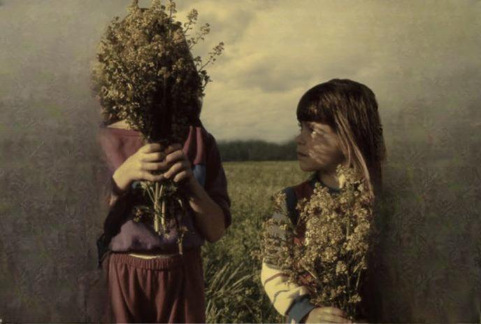 Two kids in a grassy field hold wildflowers; one hides their face while the other smiles softly.