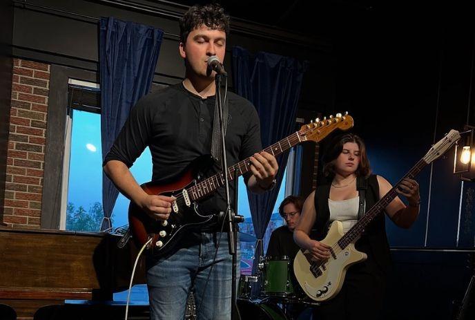 Three musicians performing on a dimly lit stage: one plays an electric guitar, another with a bass guitar, and a drummer.