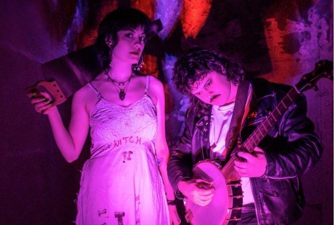 Two artists in purple lighting; one holds a banjo, and the other one is wearing a witch dress.