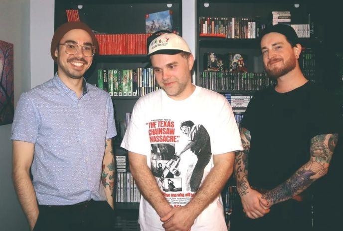 Three tattooed artists stand near shelves with books and games posing for a picture.
