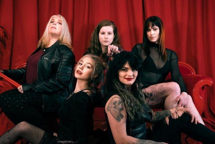 Five artists pose on a red couch before a red curtain, dressed stylishly and smiling for the camera.