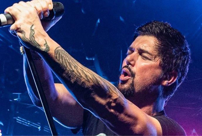 A tattooed person holding a mic on stage, bathed in vivid blue and purple light, performing passionately.
