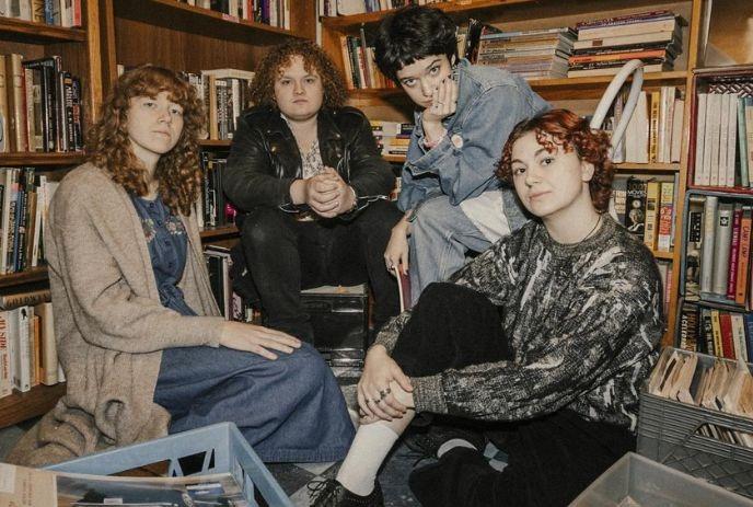Four band members sitting together surrounded by books and shelves, posing for a picture,