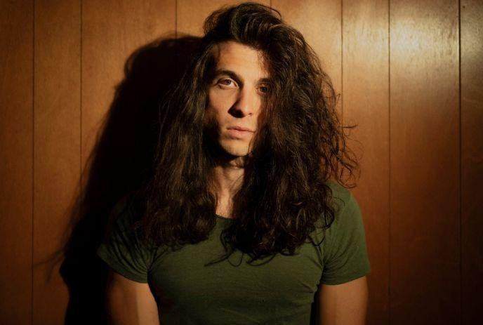 Artist with long hairs covering his face wearing an olive shirt under a wooden brown background.