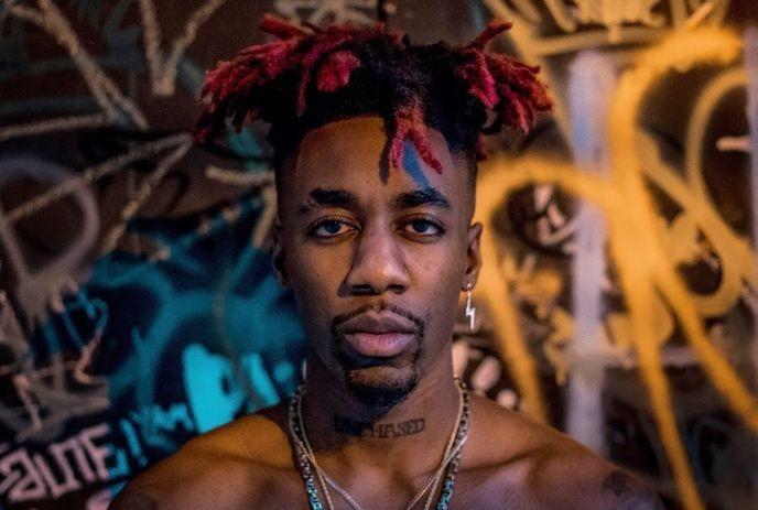 Artist with red-tipped dreadlocks stands in front of a vibrant graffiti wall with tattoos and layered necklaces.
