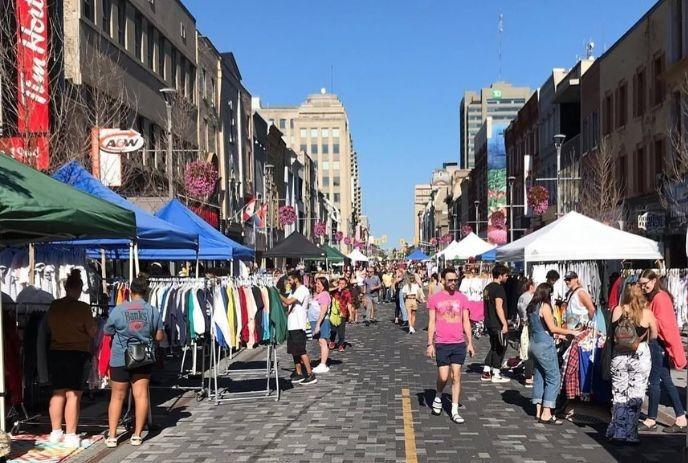 Sunny street market with stalls selling clothes, people shopping, walking, buildings in background. Vibrant scene.