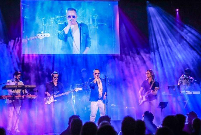 Stage lit in purple and blue with band members playing guitar, keyboard, and singing in front of crowd.