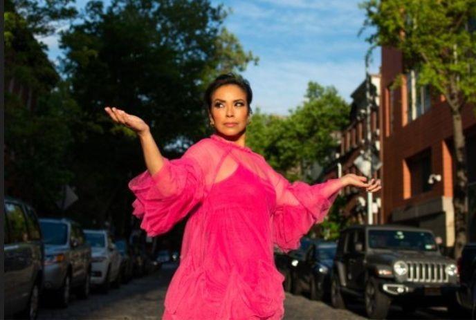 Emm Gryner in a vivid pink dress raises arms joyfully on a cobblestone street, framed by parked cars and leafy trees.
