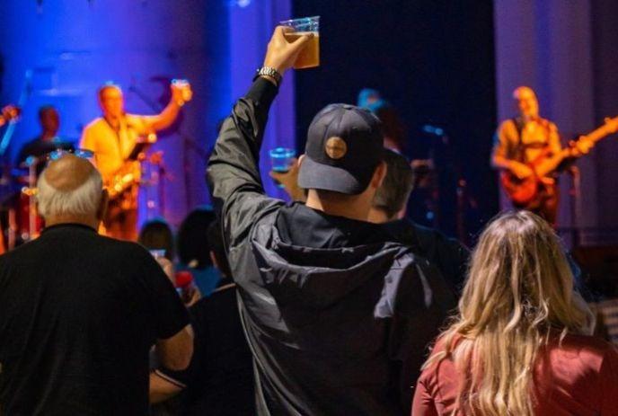 Crowd cheers as band plays under vibrant lights; a fan in black raises a drink, soaking in the concert vibe.