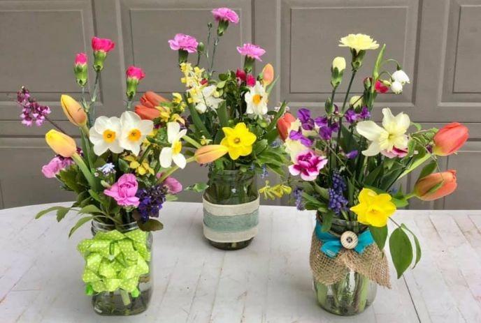 Three jars with spring flowers, each wrapped in ribbon or burlap, sit on a white surface before gray wall.