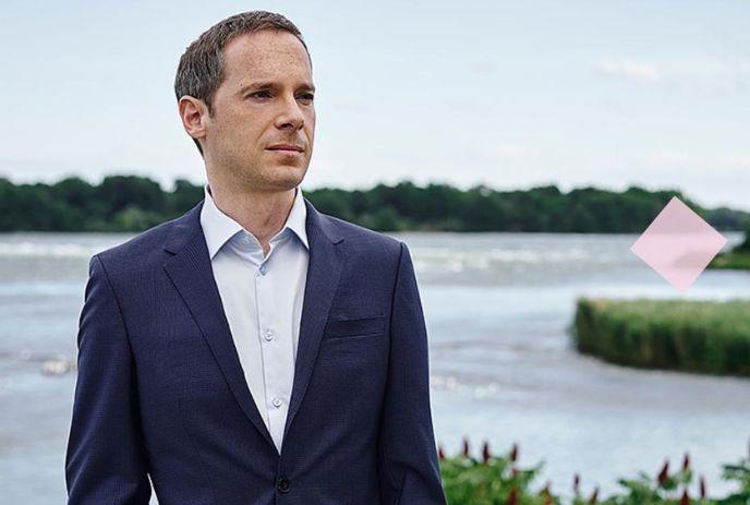 Artist in blue suit stands by water with trees in background with clear sky.