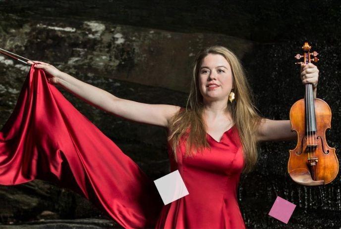 Woman artist in red dress holds violin and bow, standing confidently against a dark, textured background.