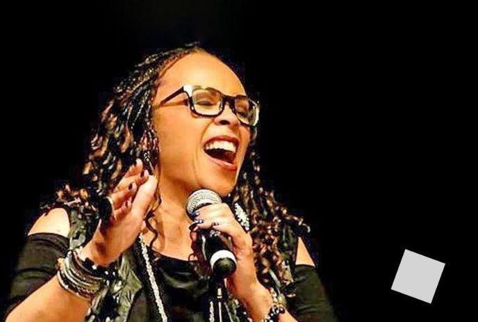 Woman artist in black outfit passionately sings on stage, holding a mic, with braided hair and bracelets, dark backdrop.