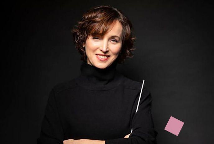 Lady in black turtleneck smiling against dark backdrop with floating pink square nearby.
