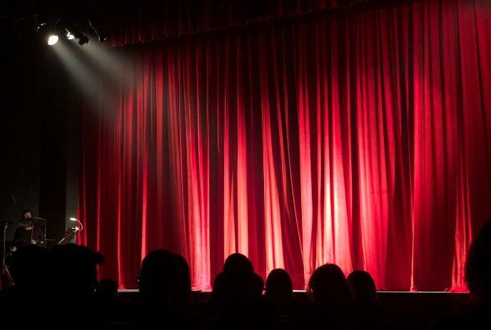 Red stage curtain glows under spotlight; audience silhouettes watch, awaiting the start of a performance.