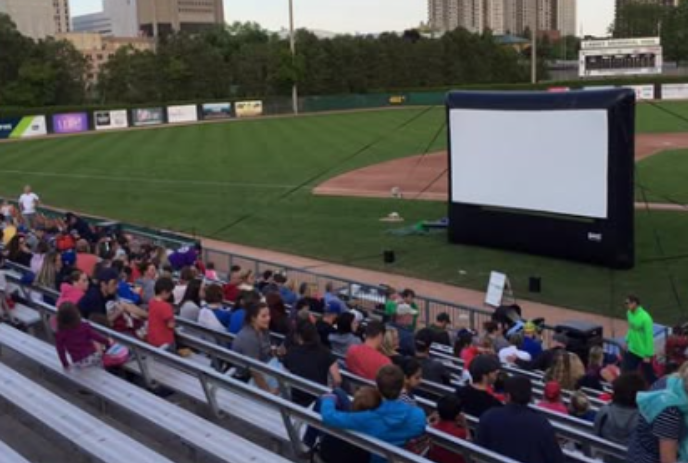 People sitting in the baseball stands watching a large movie screen on the pitcher's mound.