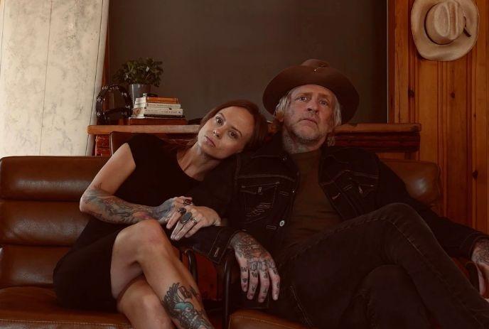 Tattooed artist duo sits close on couch in warm wood room, posing for a picture.