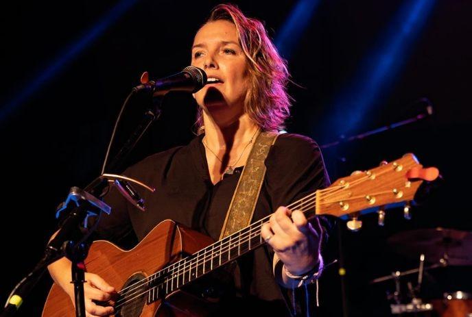 Singer strums acoustic guitar under moody lights, harmonica ready—live music moment.