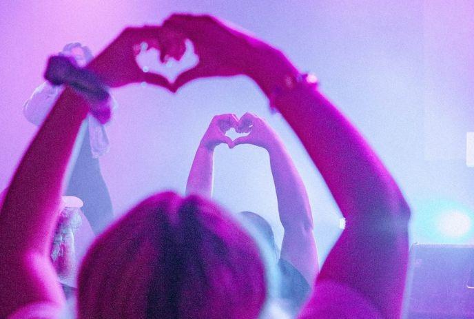 People at a concert making heart shapes with hands under pink and purple lights, showing love.