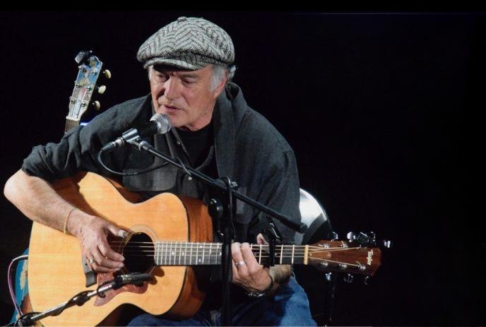 Artist in flat cap playing acoustic guitar and sings on stage, spotlighted against a dark backdrop.