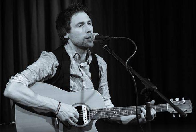 Artist sings into mic while playing acoustic guitar on stage; black-and-white, expressive live performance.