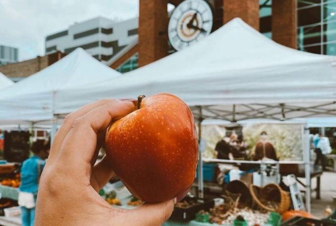 Hand holding a red apple at a sunny farmers market with white tents, shoppers at the Covent Garden Market.