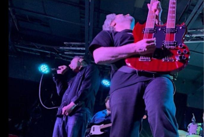 Musician plays red double-neck guitar on stage as singer performs nearby, lit by dramatic blue and purple concert lights.
