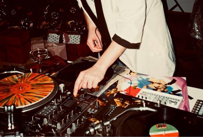 DJ in festive setting spins colorful vinyls; Christmas tree, gifts, and wine add to the party vibe.