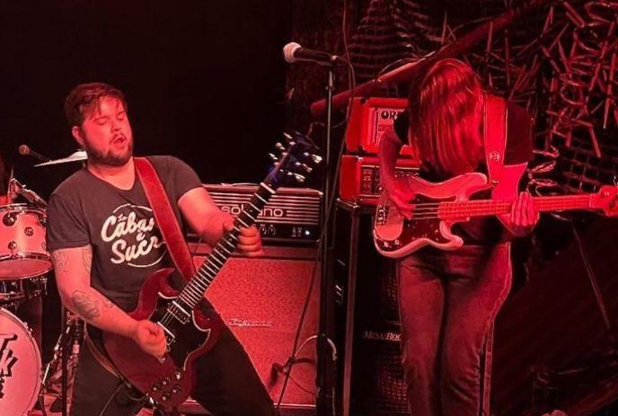 Two musicians perform on stage—one with guitar, one with bass—red lighting, tattoos, long hair, intense vibe.