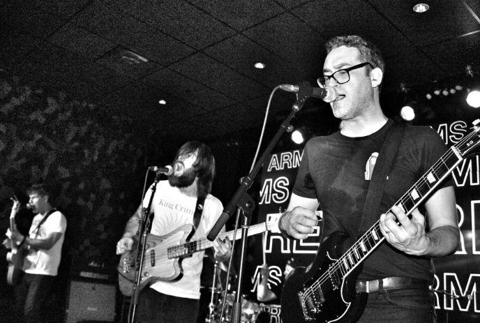 Three musicians perform live in black and white—two on electric guitar, one on bass—each singing with intensity.