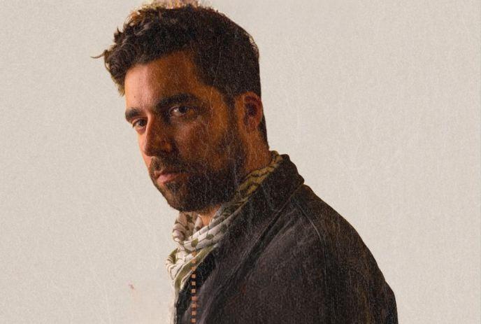 Man with beard and scarf gazes sideways, lit softly against pale backdrop—quiet, thoughtful presence.