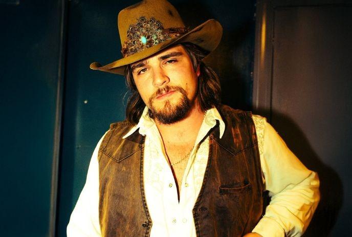 Person in a brown cowboy hat, white embroidered shirt, and suede vest, standing indoors against a dark background.
