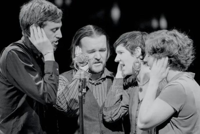 Four singers stand close around one microphone, hands over ears, harmonizing on dim stage; black-and-white photo.