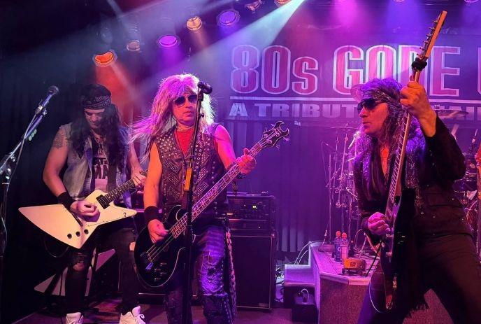 Three rock musicians in 80s attire play guitars on stage lit with red, purple, and orange lights.
