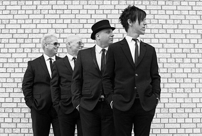Four artists in black suits pose by white brick wall facing left in black-and-white style.