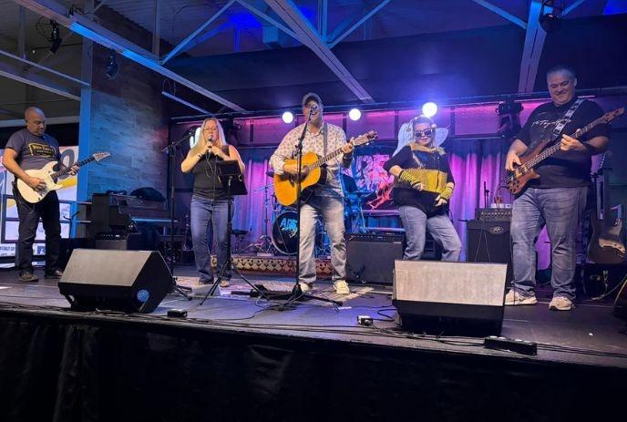 Five musicians perform on lit stage with guitars, mics, and energy; purple-blue lights and logo backdrop.